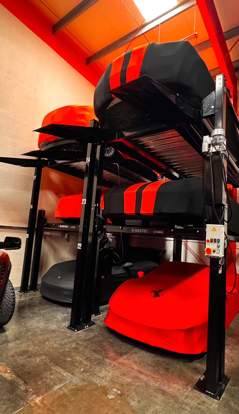 Three-level car lift with covered cars and a Ferrari under a red cover